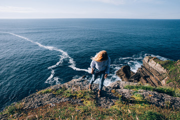 Blonde pretty woman in jeans and shirt looks at amazing ocean scenery, waves and cliffs, wind plays with her hair and she dreams about future and peace
