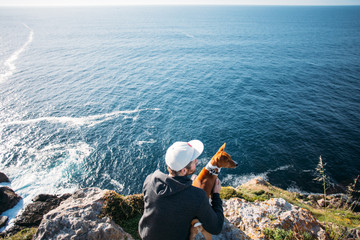 Hipster millennial young man in hoodie sweatshirt and five panel cap sits on cliff and overlooks sunset over ocean together with best friend, basenji dog - Powered by Adobe