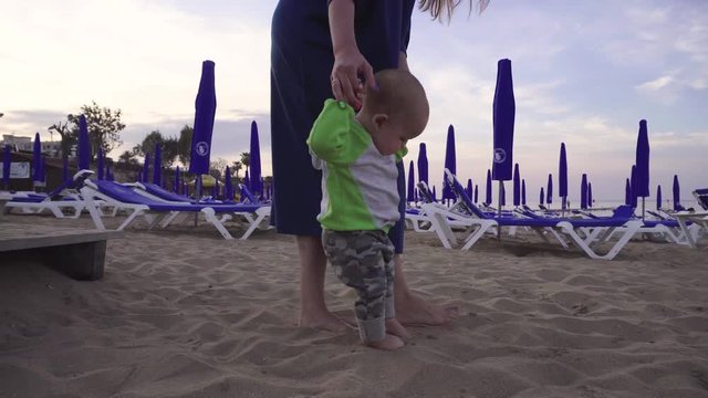 Joyful little boy makes first steps on a sandy beach holding mother's hands. Handheld shot
