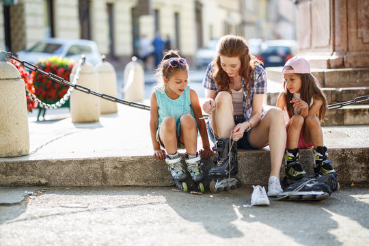 Mother plays with her daughters on street in neighborhood.They sitting on stairs in city square and preparing to drive a  roller blades .Family concept.