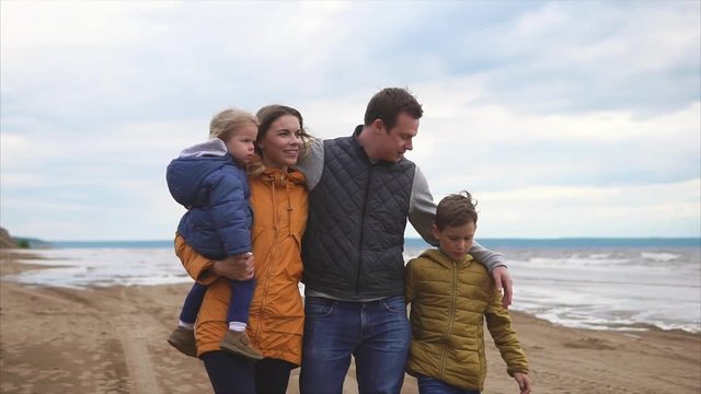 A happy and friendly family is walking with their young children on the beach near the sea on a day off, mom is carrying a baby, a middle-aged man is hugging his wife and son