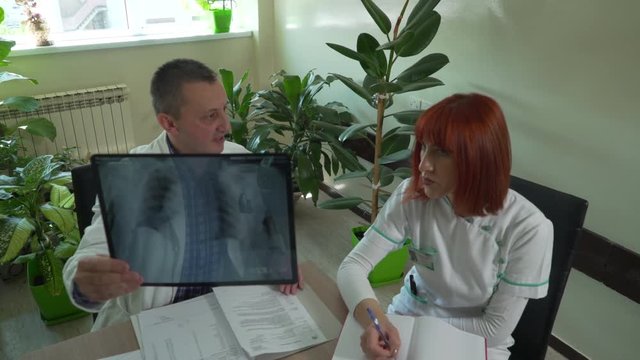 Middle aged Caucasian male doctor and nurse with red hair in white gowns sit at the table and together examine x-ray image of patient lungs on a meeting at office, crane, tracking shot, room interior