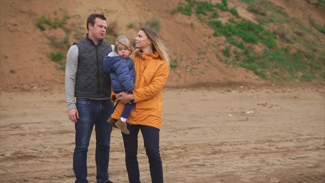 A young married couple is standing on a sandy coast, an adult mother is holding a son in her arms, her middle-aged husband is standing next to him and lovingly looks at his wife