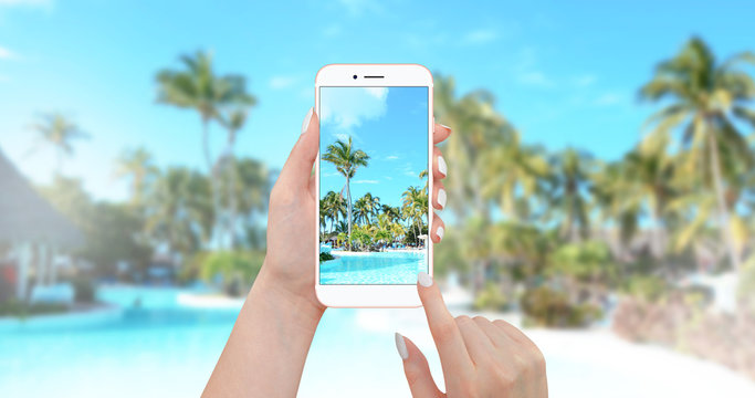 Girl taking picture of pool with smartphone with palm trees and beach in background