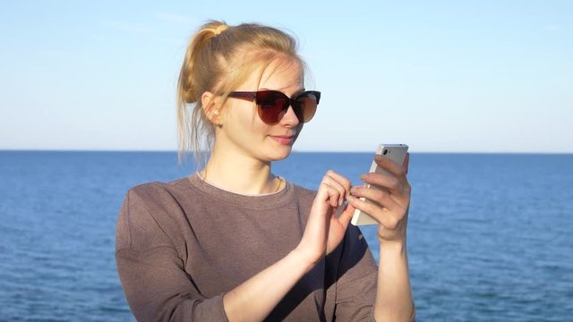 Beautiful young blonde girl in sunglasses uses a smartphone of white color near the sea. A teenager dials a number and writes a message in social networks on the phone in front of blue sea