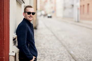Obraz premium Confident fashionable man leaning against a wall in an alley wearing a blue leather jacket and sunglasses smiling towards camera.