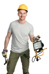Smiling man in a grey shirt and green pants with a safety helmet standing in front of a white background holding a hammer and a lamp. Looking at camera.