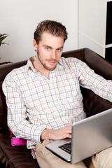 Handsome man wearing a bright shirt and pants, sitting down in a sofa indoors with a laptop...