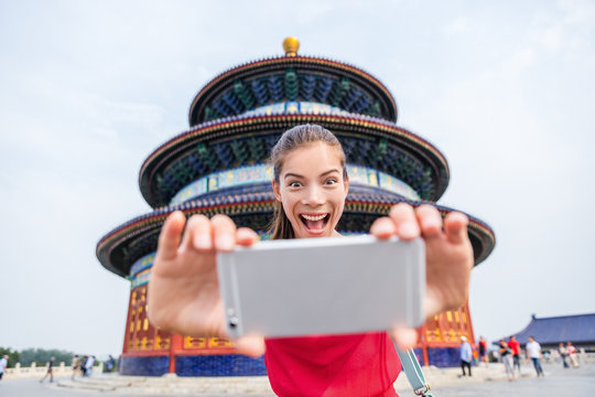 Tourist taking goofy face funny selfie on travel. Happy young woman having fun with mobile phone at famous Beijing landmark. Temple of Heaven china travel.