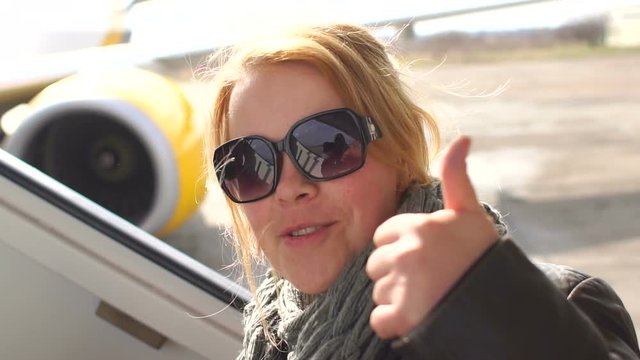 Young girl in sunglasses showing thumbs up standing next to the plane before departure.