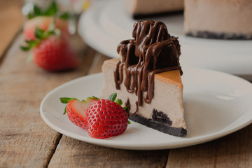 Homemade chocolate cheesecake on rustic wood table. Baked chocolate cheesecake topping brownie and dark chocolate sauce decoration with strawberry. Triangle slice piece of cheesecake on white plate.