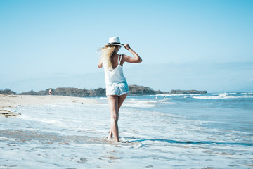 Young sexy woman with white hat walking on white sand beach a tropical Bali island at sunny day. Ocean cost.