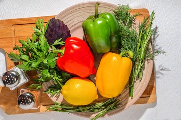Fresh bright vegetables: Bulgarian pepper, herbs, lemon, on a ceramic plate, on a wooden cutting board