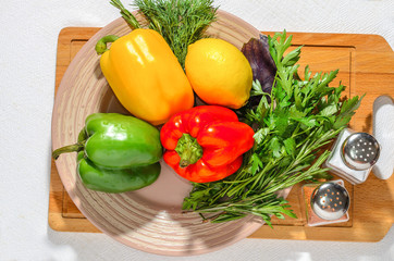 Fresh bright vegetables: Bulgarian pepper, herbs, lemon, on a ceramic plate, on a wooden cutting board