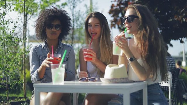 Group of three young trendy women sitting at small table in outside cafe having refreshing drinks in hot summer day