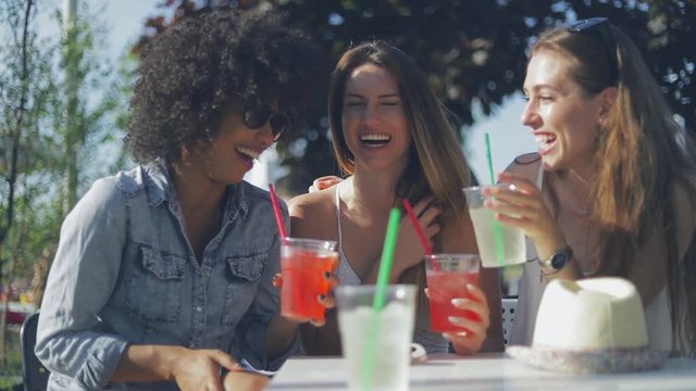 Three young women in summer clothing sitting at table with drinks in outside cafe and laughing while having fun