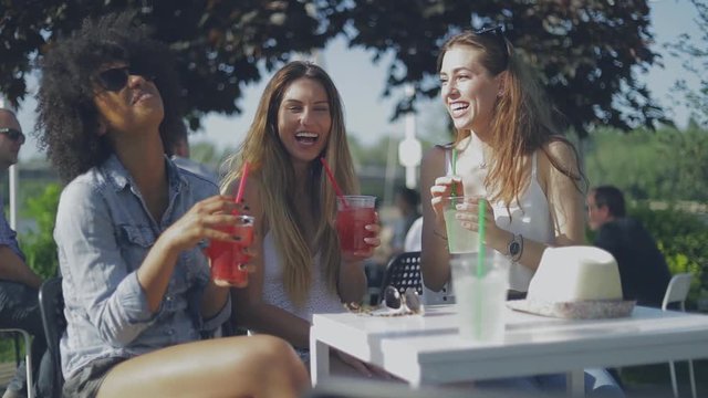 Three young girls in stylish summer clothing enjoying freshly made drinks while sitting at table in cafe and laughing