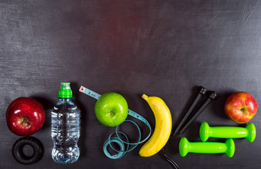 Dumbbell, jump rope, apple, banana and water bottle on black background. Top view fitness equipment background
