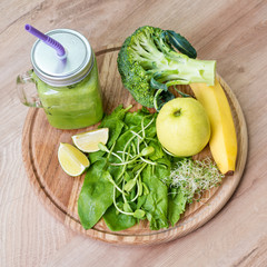 Fresh green vegetables and green smoothie in jar. Detox, diet or healthy food concept. Mason jar of dietary drink with broccoli, spinach, microgreens, lime and banana on wooden background. Top view.