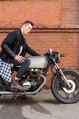 Naklejka na ściany i meble Handsome rider biker guy in leather jacket sit on classic style cafe racer motorcycle. Side view. Vertical photo. Bike custom made in vintage garage. Brutal fun urban lifestyle. Outdoor portrait.