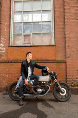 Fototapeta na wymiar Handsome biker guy in black leather jacket sit on classic style cafe racer motorcycle and hold helmet on gas tank. Bike custom made in vintage garage. Brutal fun urban lifestyle. Outdoor portrait.