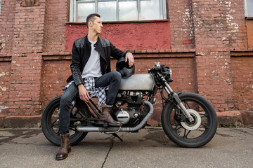 Plakat Handsome rider biker man in black leather jacket, jeans, boots and helmet sit on classic style cafe racer motorcycle. Bike custom made in vintage garage. Brutal fun urban lifestyle. Outdoor portrait.