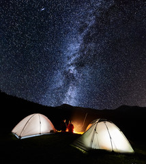 Night camping in the mountains. Rear view of romantic couple tourists have a rest at a campfire near two illuminated tents under amazing night sky full of stars and milky way. Low light