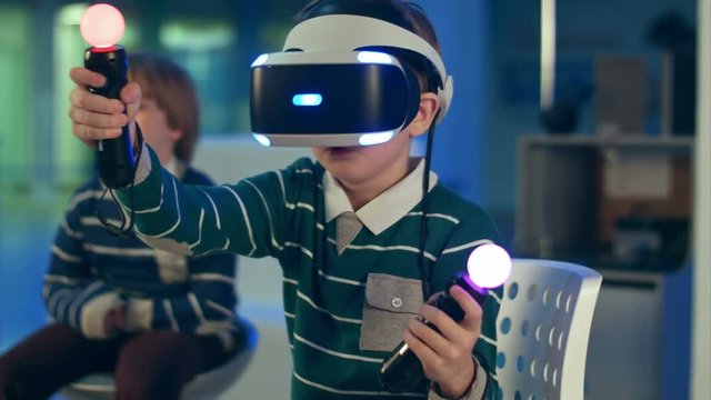 Little boy in vr headset playing virual reality game with controllers while another boy waiting for his turn