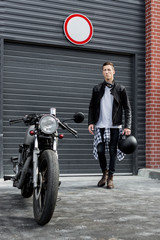 Beautiful young rider man in black biker jacket and boots go to his classic style cafe racer motorcycle industrial gates as background. Bike custom made in vintage garage. Brutal fun urban lifestyle.