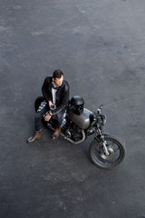 Plakat Top view of a handsome rider guy in black biker jacket and check shirt look to smartphone phone on classic style cafe racer motorcycle. Bike custom made in vintage garage. Brutal fun urban lifestyle.