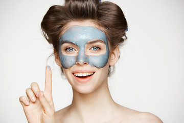 Portrait of young tender girl in hair curlers and facial mask looking at camera smiling over white background. Beauty and skincare concept.