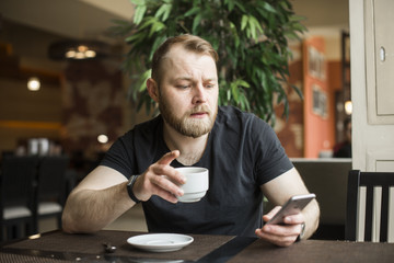 Fototapeta na wymiar Young bearded businessman,dressed in gray cardigan,sitting at table in cafe and use smartphone. Man holding smartphone and looking at its screen. Man using gadget. Guy browsing internet on smartphone