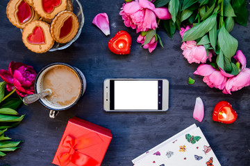 Love and care concept. Romantic style composition - smart phone surrounded with peonies, cookies and mug with coffee, candles, present box on the black wooden background. Top view. space for text.