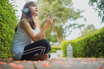 Young happy smiling asian woman listening music with headphones from her smart phone at the park during a sunny day wearing white clothes	