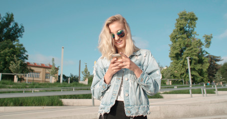 Young blonde woman using phone in a city. Beautiful young woman typing on phone during sunny day.