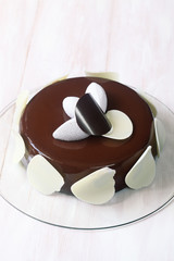Contemporary Chocolate Coconut Mousse Cake covered with chocolate mirror glaze, on transparent glass board, on white wooden background.