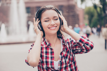 Funky spring mood! Chilling outside. Young happy lady is listening to the music outdoors, wearing big white modern earphones and checkered shirt with closed eyes