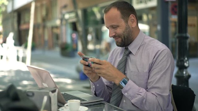 Happy businessman playing game on smartphone sitting in cafe in city
