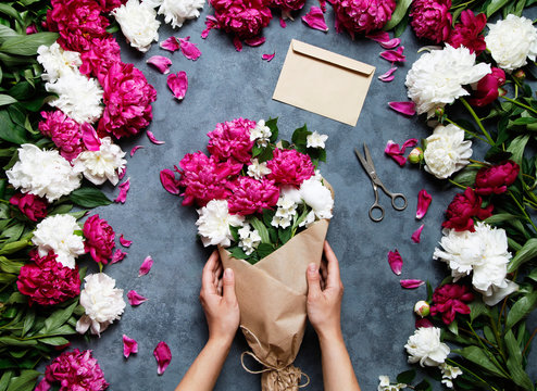Female florist holding beautiful bouquet at flower shop. Florist at work: pretty woman making summer bouquet of peonies on a working gray table. Kraft paper, scissors, envelope for congratulations.