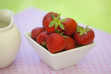 Strawberries in a white angled bowl on a pink dotted tablecloth with green bokeh in the background