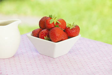 Strawberries in a white angled bowl on a pink dotted tablecloth with green bokeh in the background