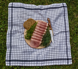 Smoked ham,basil,dill,bread and knife on a wooden plate on table clouth on the grass