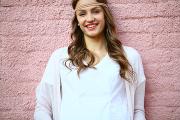 Positive hipster woman with wicker headband standing on pink brick wall, looking camera