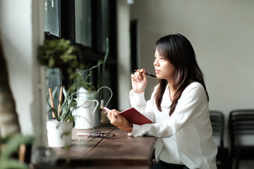 young women holding pen for writing on notebook. her sitting by the window at library room in morning. image for education,business,people,interior and portrait concept