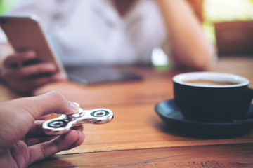 A hand holding and playing metal silver color fidget spinner with coffee cups and people using mobile phone in background