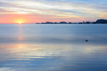 Naklejka premium Beautiful seascape view when sunset at Nang Rum beach located in Chon Buri in eastern of Thailand with port, hill, lighthouse, cloud and twilight sky as background. Complementary color. Long exposure.