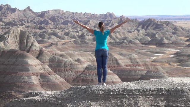 SLOW MOTION: Young woman trekking Badlands National park, standing on top of rocky mountain in with her arms raised up towards sunny sky enjoying and relaxing. Traveler exploring Badlands wilderness