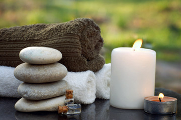 Fototapeta na wymiar Spa still life with towels, a burning candle, bath oil and massage stones against the backdrop of a green garden in summer
