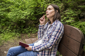 Beautiful woman sitting on a wooden bench in the forest park at the trail and dreamily looks up at the sky, carrying a notebook and a pencil in his mouth.