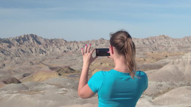 CLOSE UP: Cheerful young woman traveler taking photos of stunning Badlands National Park on a sunny day in South Dakota, USA. Happy girl exploring desert wilderness and photographing with mobile phone